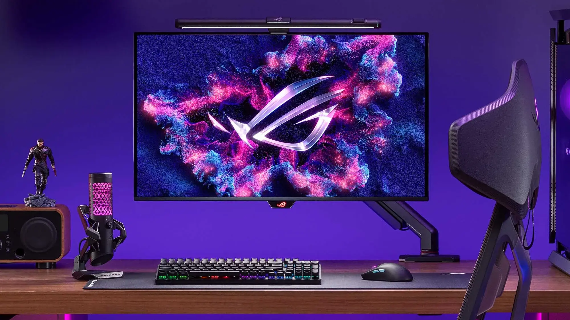 A gaming setup featuring an ASUS ROG Xreal R1 augmented reality device mounted above a large monitor with the ROG logo, accompanied by a keyboard, mouse, a speaker, a purple-lit microphone, and a figurine on a wooden desk against a purple wall.