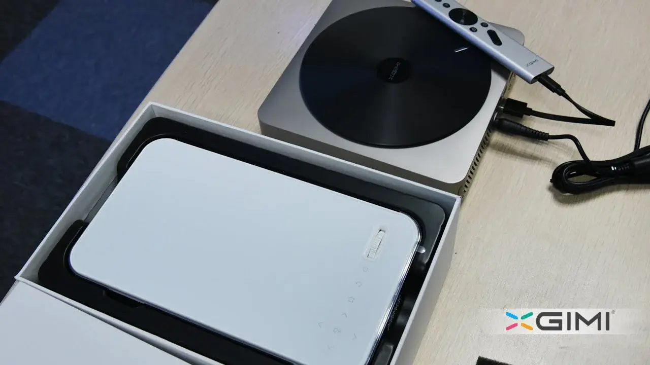 A compact, rectangular device with a sleek white top is placed inside a storage box, alongside a projector connected to cables, with the GIMI logo visible on the bottom right corner.